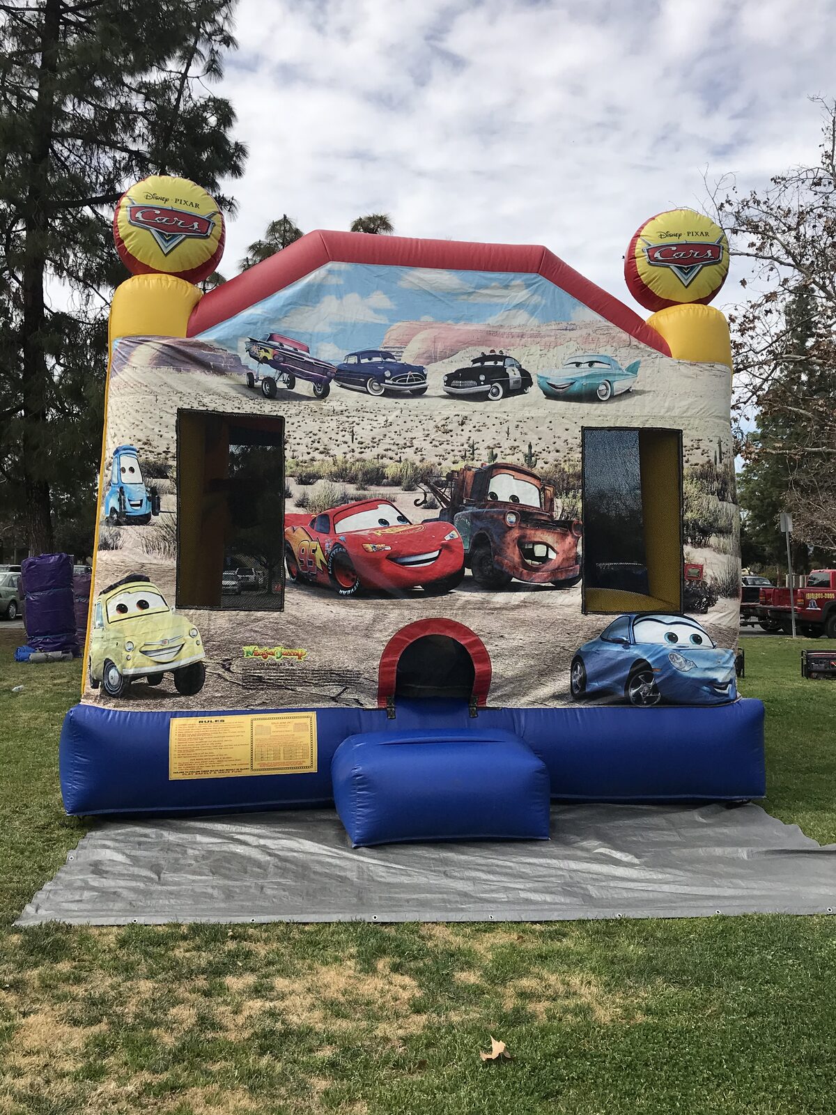 4n1 cars combo bounce house rental in San Diego, front view of the Cars-themed unit with race car art and mesh windows set up on a tarp at a park