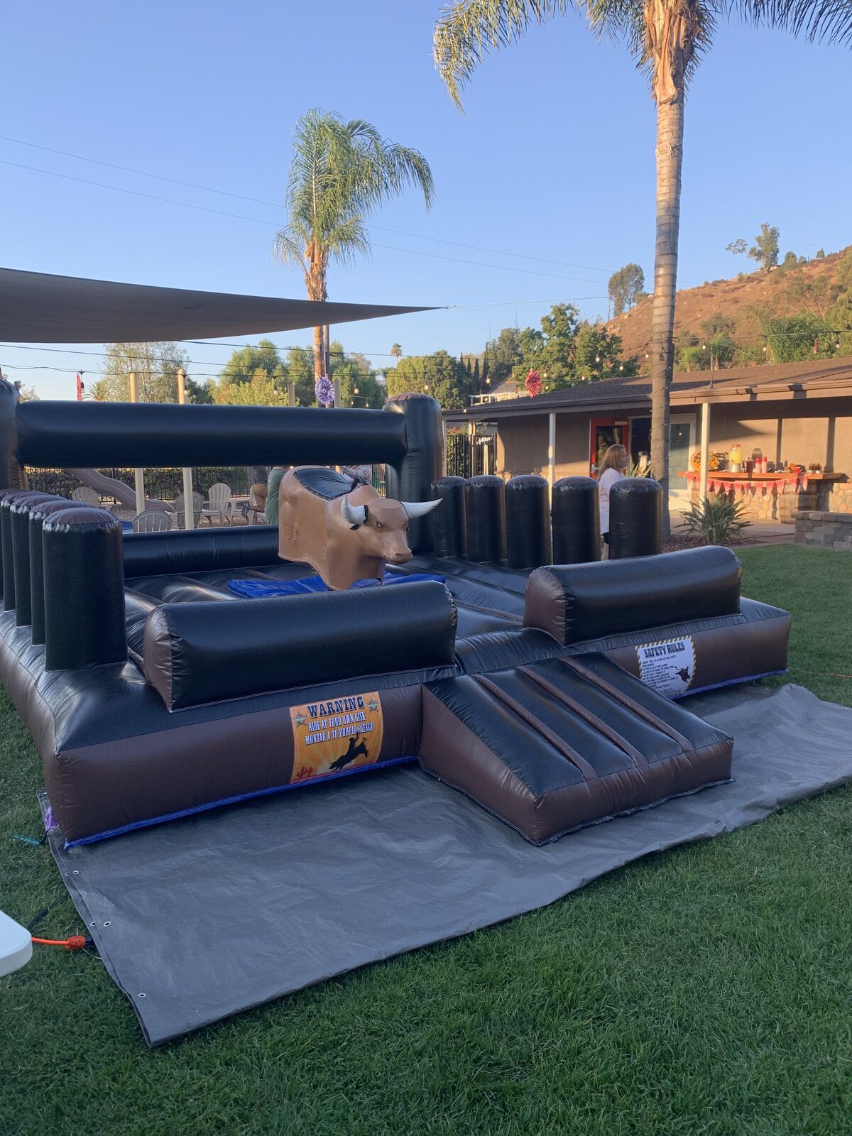 Mechanical bull rental in San Diego, 13x13 setup on grass with palm trees, showing the padded landing bumpers