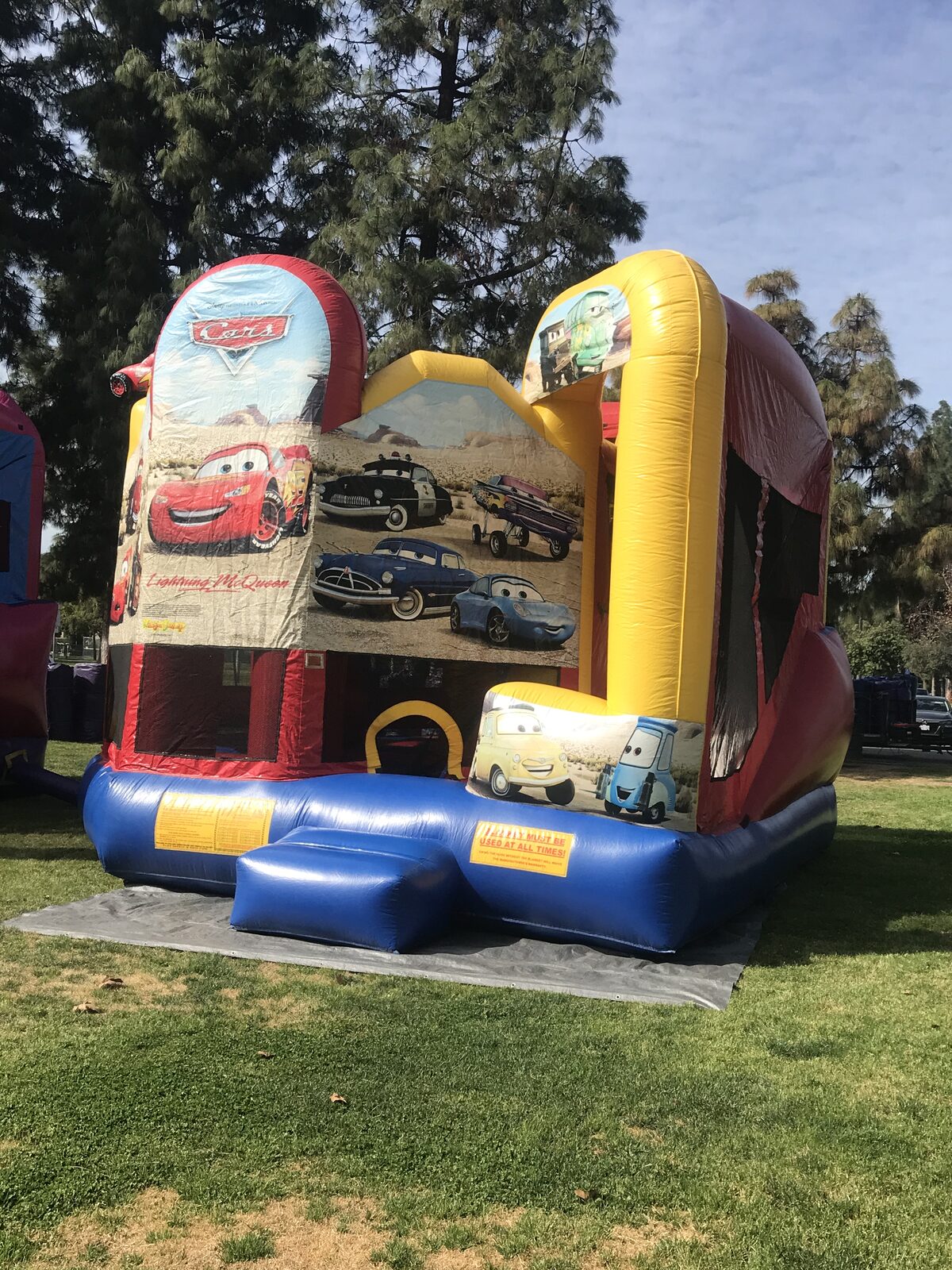 5n1 cars combo bounce house rental in San Diego, three-quarter view showing the Cars art panel, entry step and side slide tower under shade trees