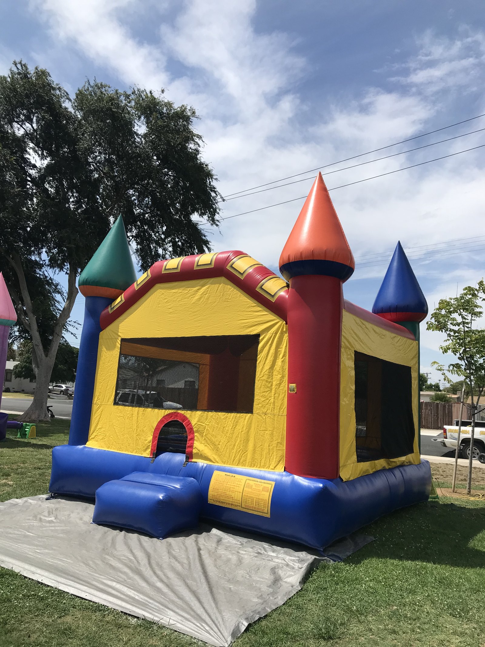 15x15 Multi-Color Castle bounce house rental in San Diego — angled view on grass over a protective tarp showing the entry ramp, rules panel, and mesh side windows