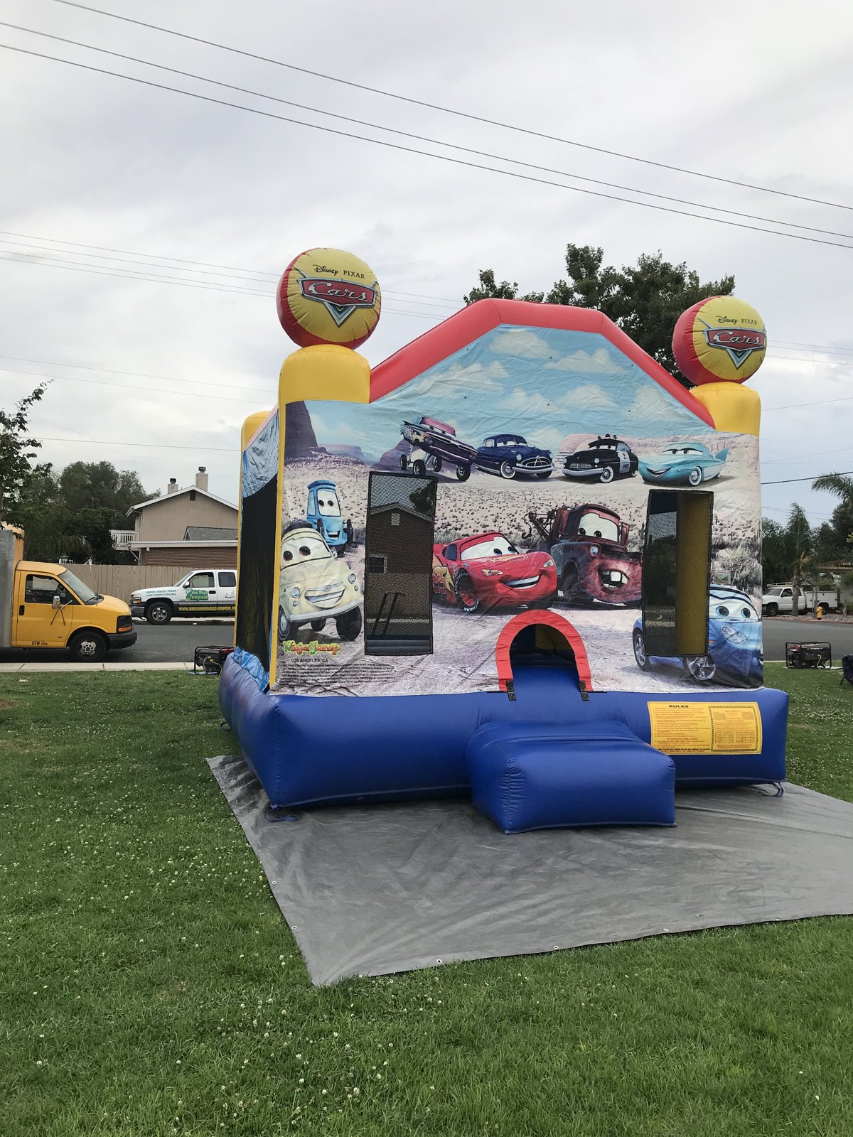 13x13 Disney's Cars bounce house rental in San Diego — angled front view with Lightning McQueen and Mater artwork and inflatable Cars logo toppers on a grass lawn