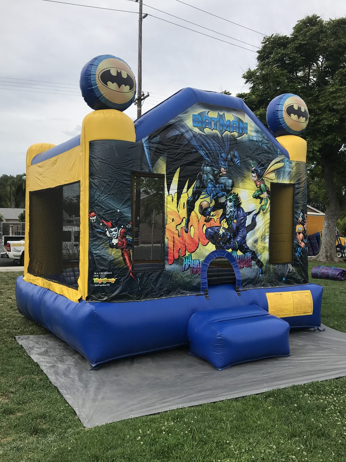 13x13 Batman bounce house rental in San Diego — angled front view on a grass lawn over a protective tarp showing the entry ramp