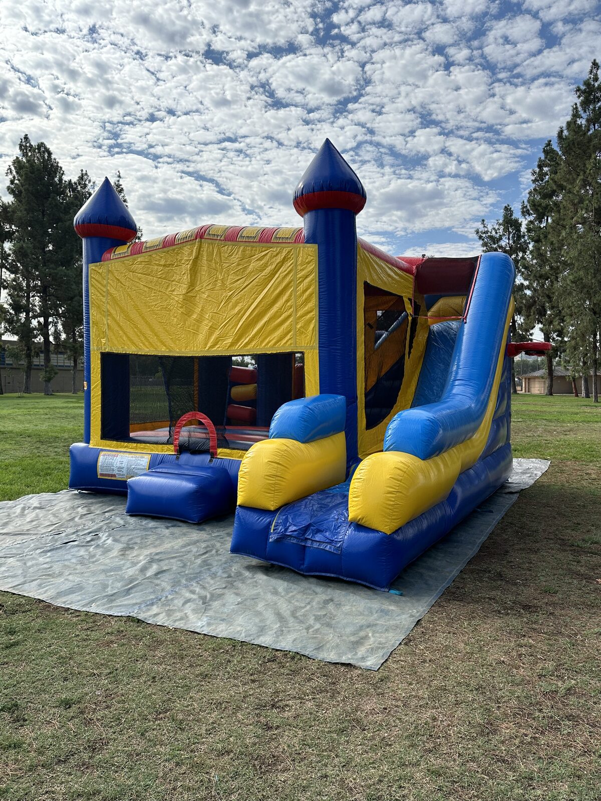 6n1 blue castle banner combo bounce house rental in San Diego, blue, yellow and red castle with turrets and a tall side slide set up on a tarp at a park