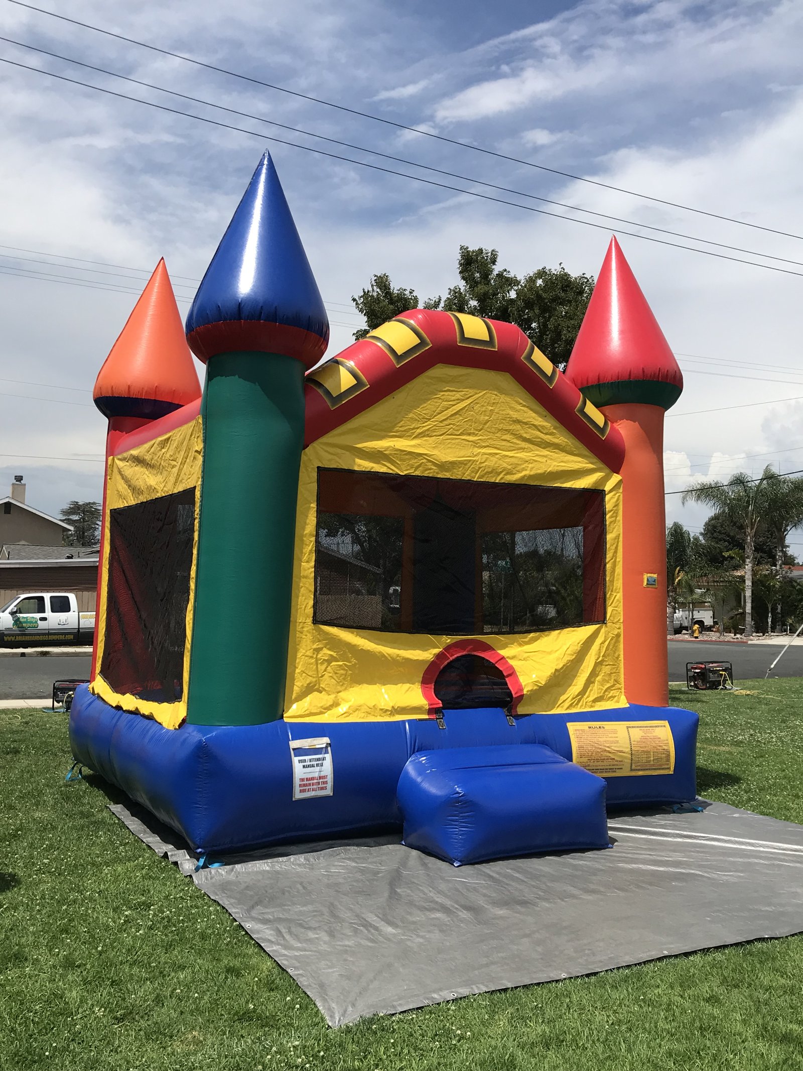13x13 Multi-Color Castle bounce house rental in San Diego — angled view on grass over a protective tarp showing the mesh side windows and blue base