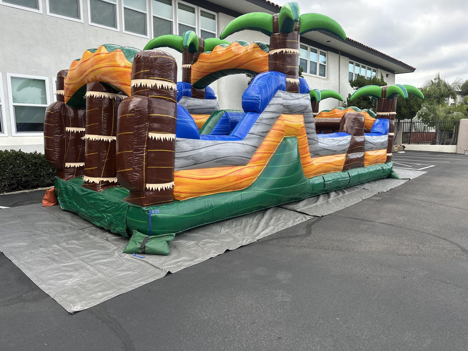 30ft tropical obstacle course rental in San Diego, three-quarter view showing the climb wall, rock-face obstacles, and palm-tree columns