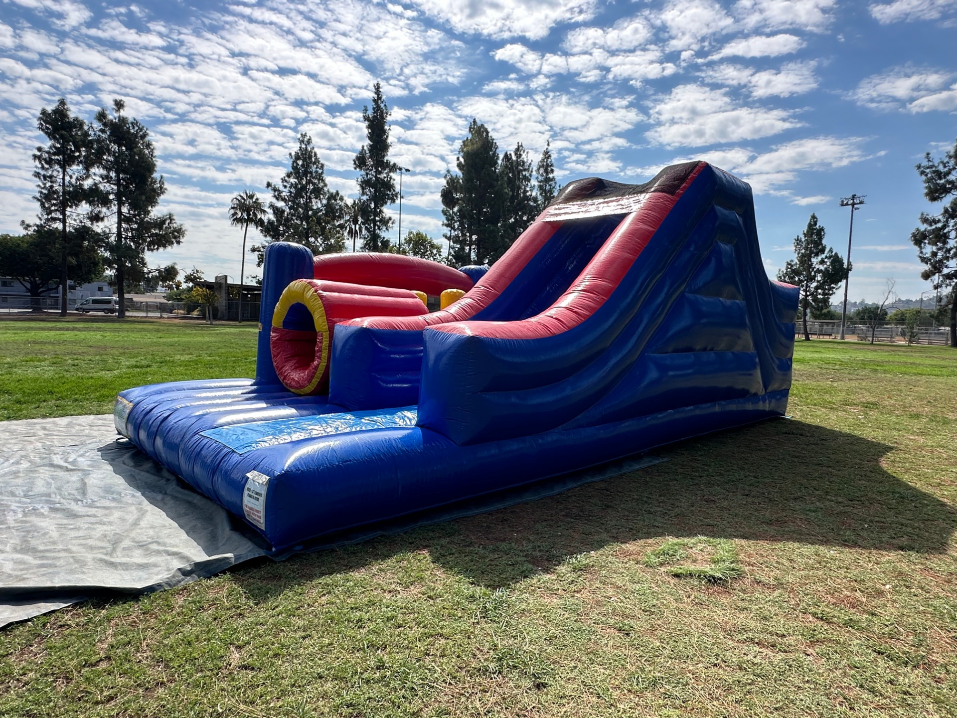 Challenge course obstacle rental in San Diego, blue and red lane, angled view of the yellow crawl tunnel and tall slide exit on a park lawn