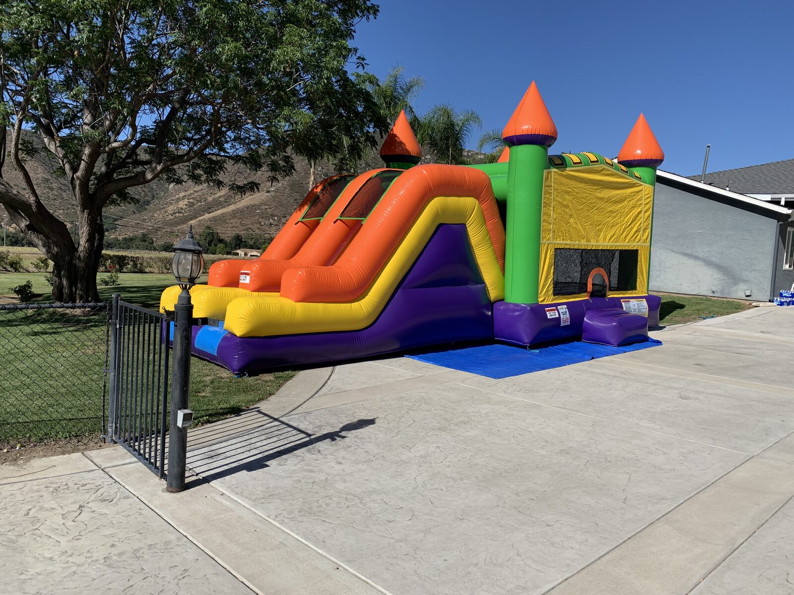 4n1 multi-color castle banner combo bounce house rental in San Diego, three-quarter view of the dual slide lanes and entry step set up on concrete beside a home