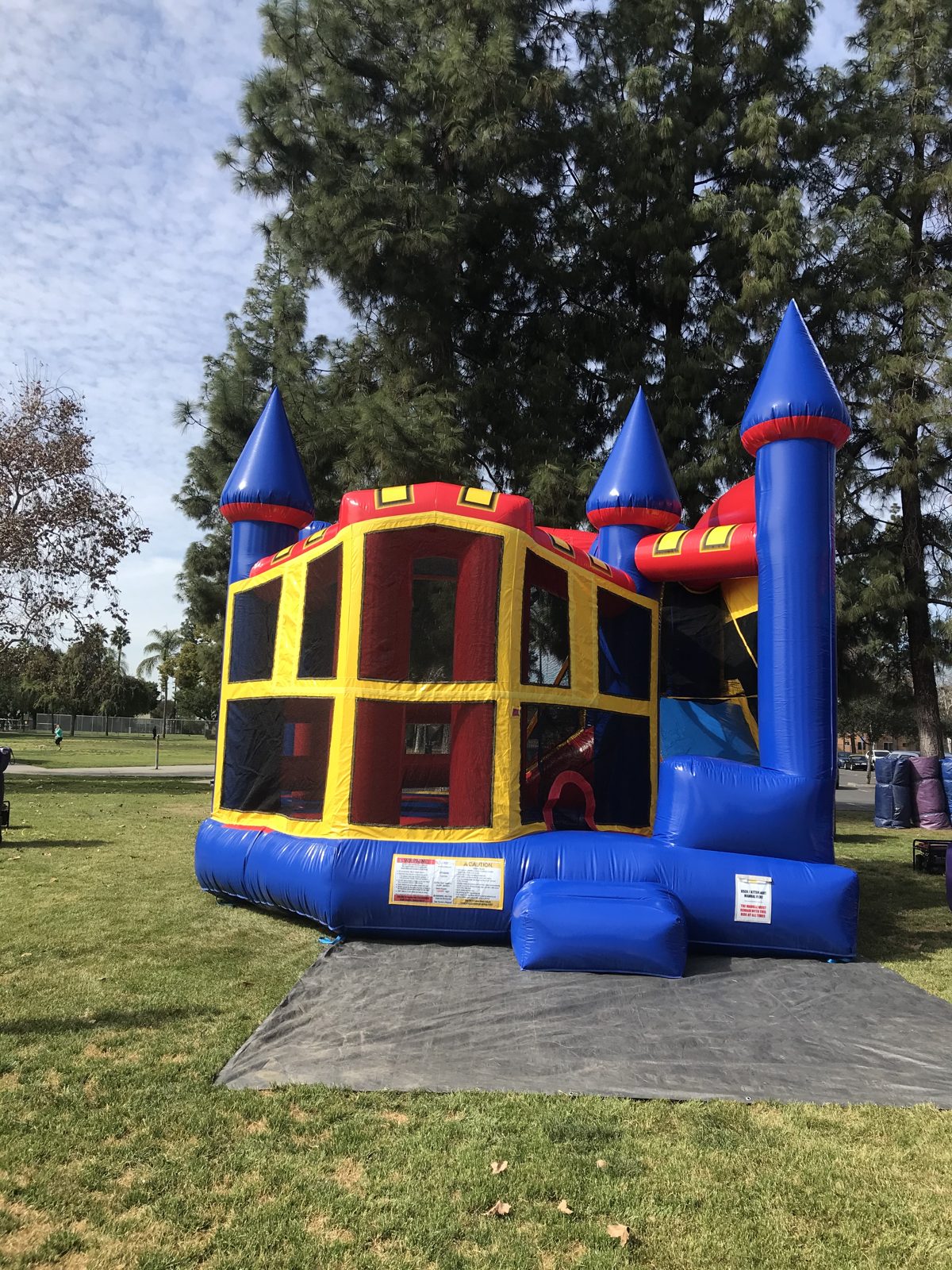 5n1 blue castle banner combo bounce house rental in San Diego, front corner view of the blue turrets and yellow mesh walls on a tarp at a park