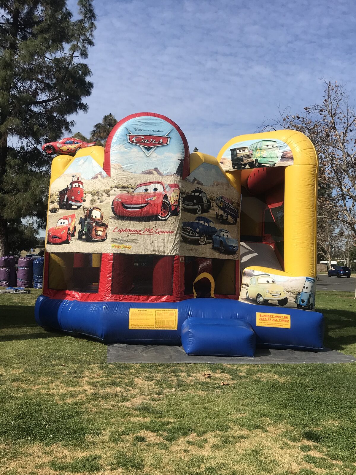 5n1 cars combo bounce house rental in San Diego, front view of the Lightning McQueen Cars themed combo with red, yellow and blue trim set up on grass at a park