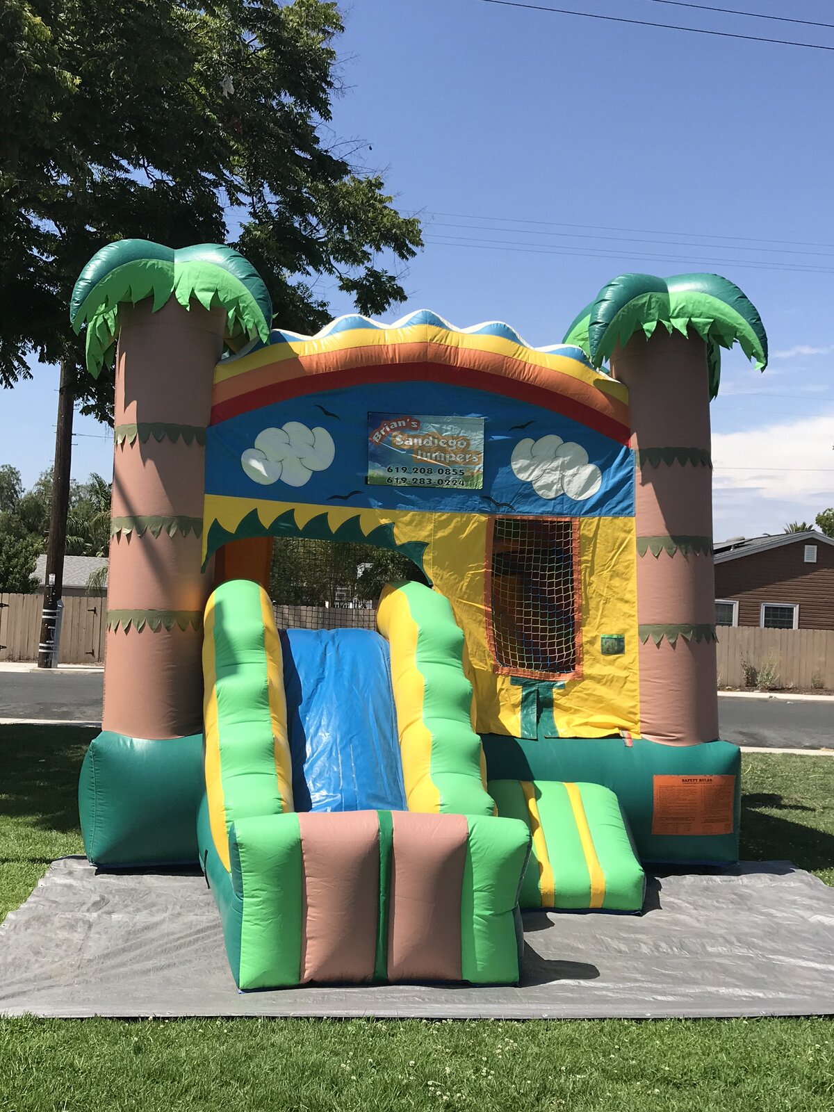 2n1 palm tree combo bounce house rental in San Diego, front view of the tropical castle with inflatable palm trees and green side slide set up on a tarp on grass