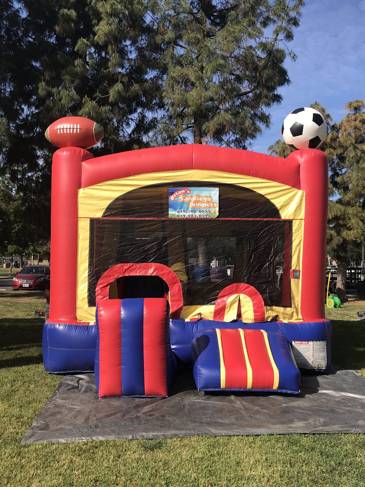 5n1 sports combo bounce house rental in San Diego, front view of the red, yellow, and blue unit with inflatable football and soccer ball toppers and step-up entry on a tarp