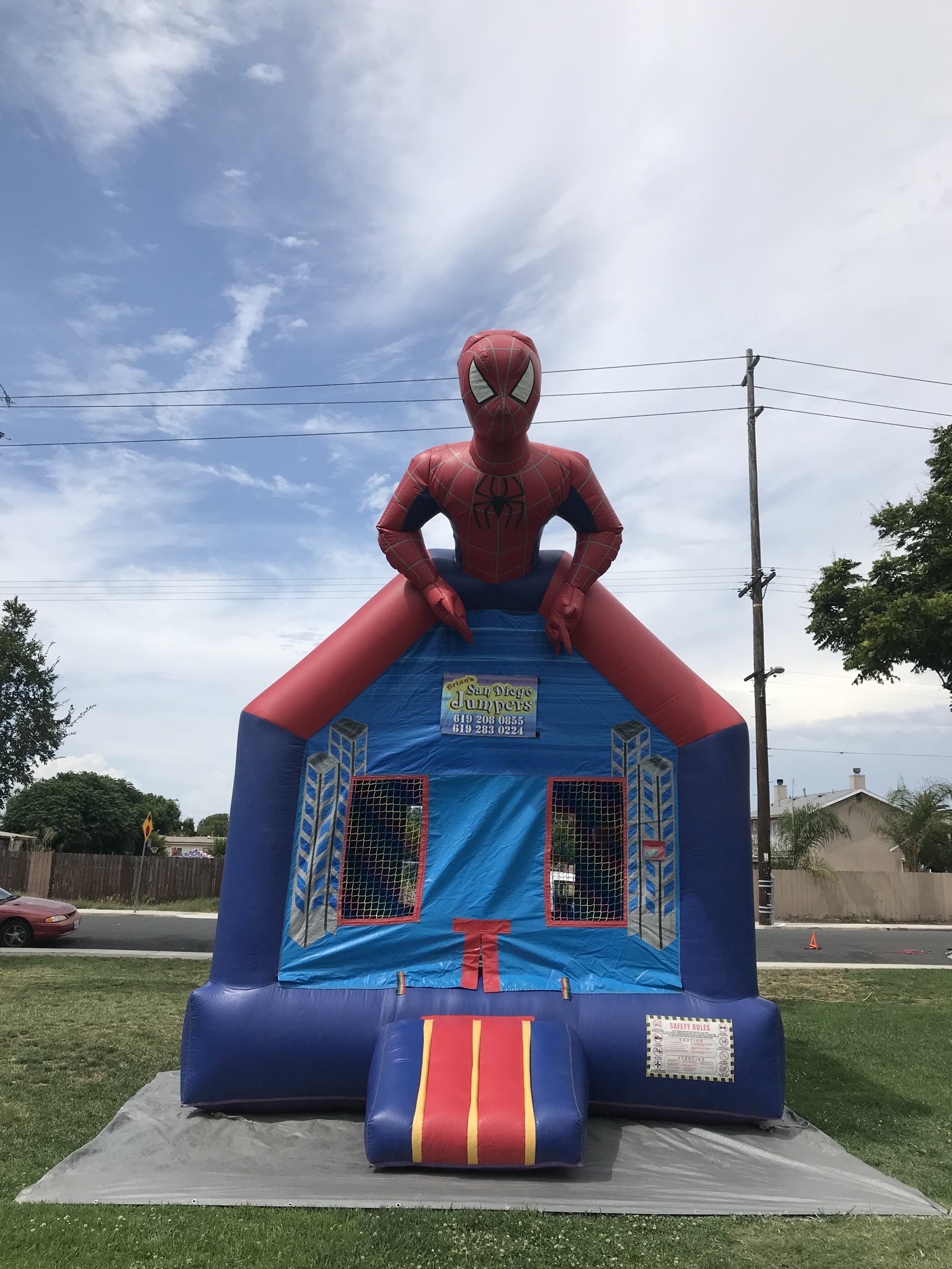 13x13 Spider-Man bounce house rental in San Diego — straight-on front view with the oversized Spider-Man topper and city skyline artwork on a grass lawn