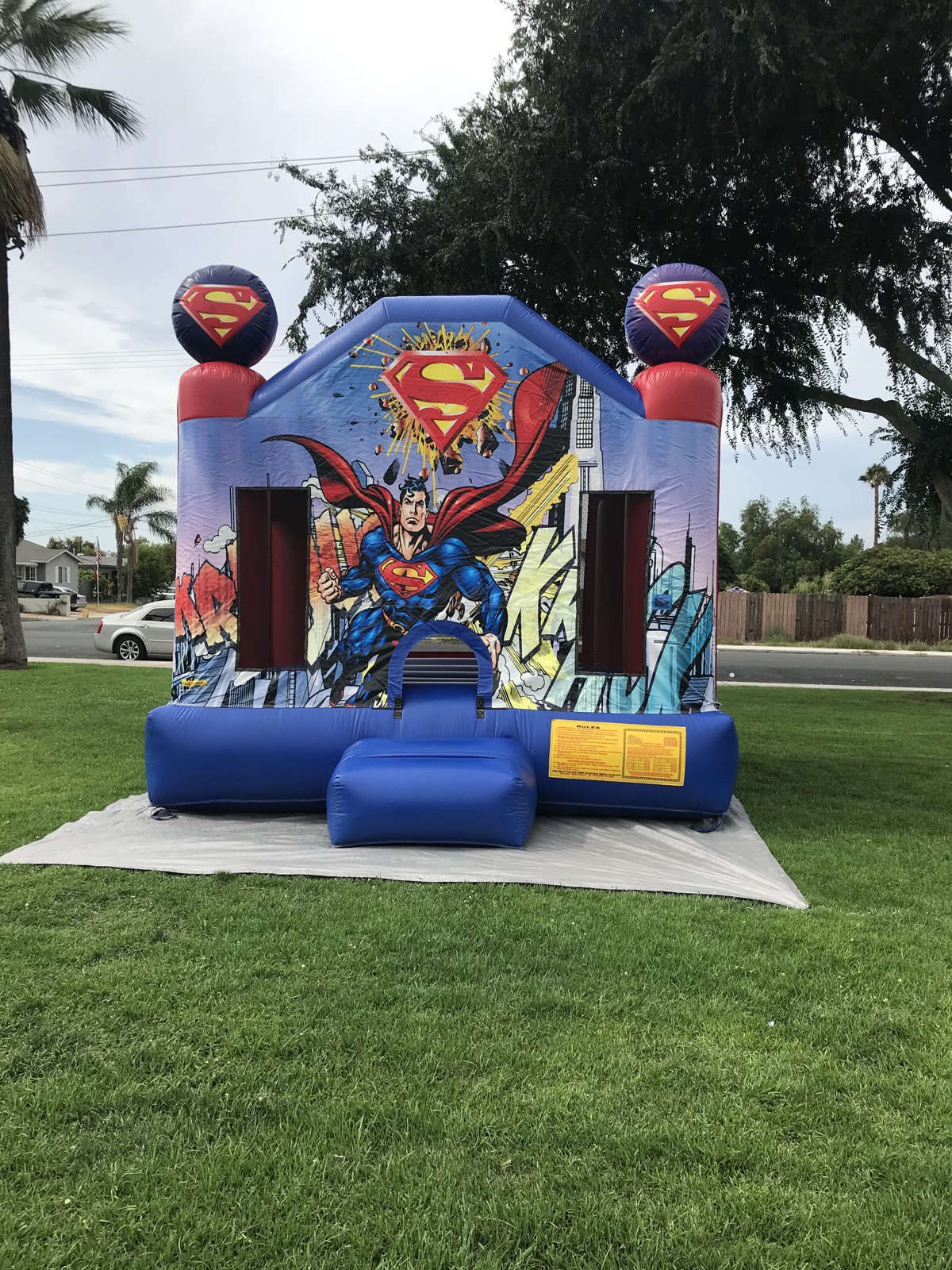 13x13 Superman bounce house rental in San Diego — straight-on front view with comic-book Superman artwork and twin Superman logo toppers on a park lawn