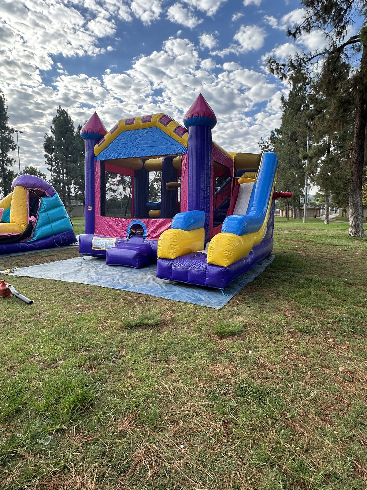 6n1 dream castle combo bounce house rental in San Diego, wide park view of the full unit staked on a tarp on grass beside a second inflatable