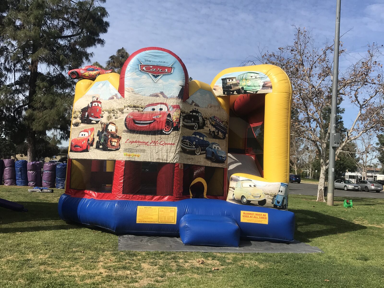 5n1 cars combo bounce house rental in San Diego, wide angled view of the full unit staked on a tarp on a park lawn with the safety rules placard visible