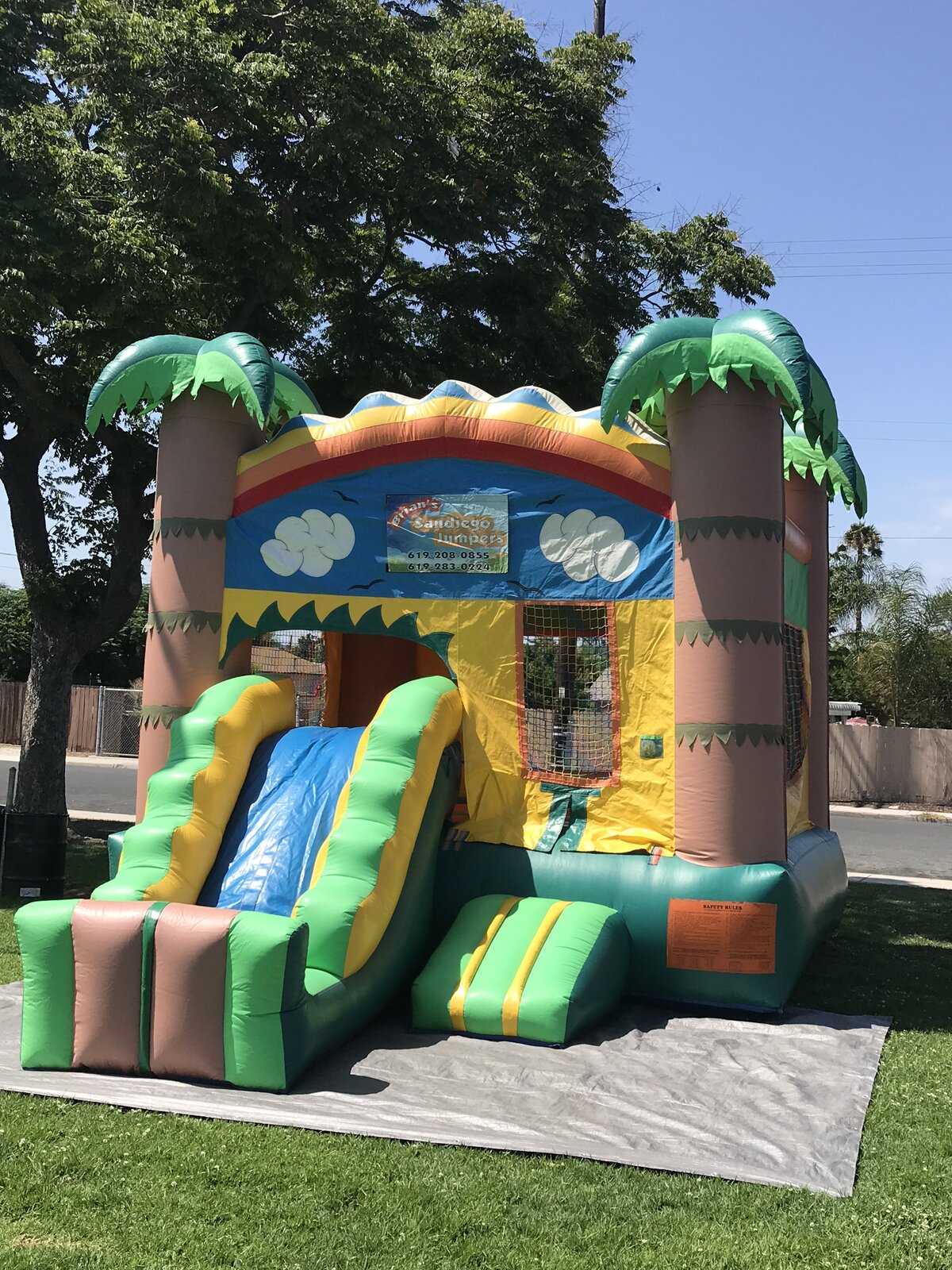 2n1 palm tree combo bounce house rental in San Diego, angled side view of the slide run and safety rules placard under shade trees