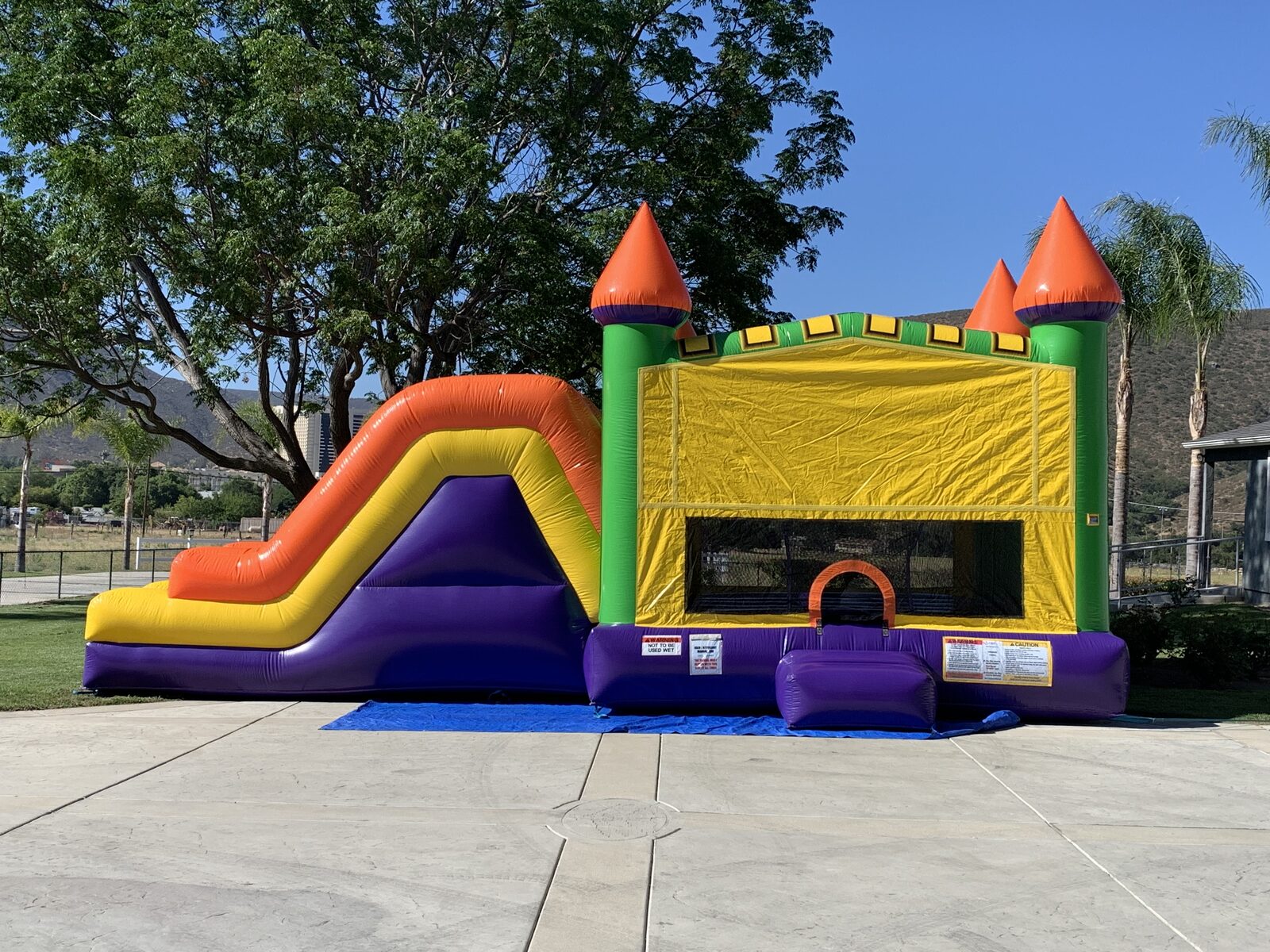 4n1 multi-color castle banner combo bounce house rental in San Diego, full side profile showing the orange and yellow slide run beside the yellow banner jump house on a driveway
