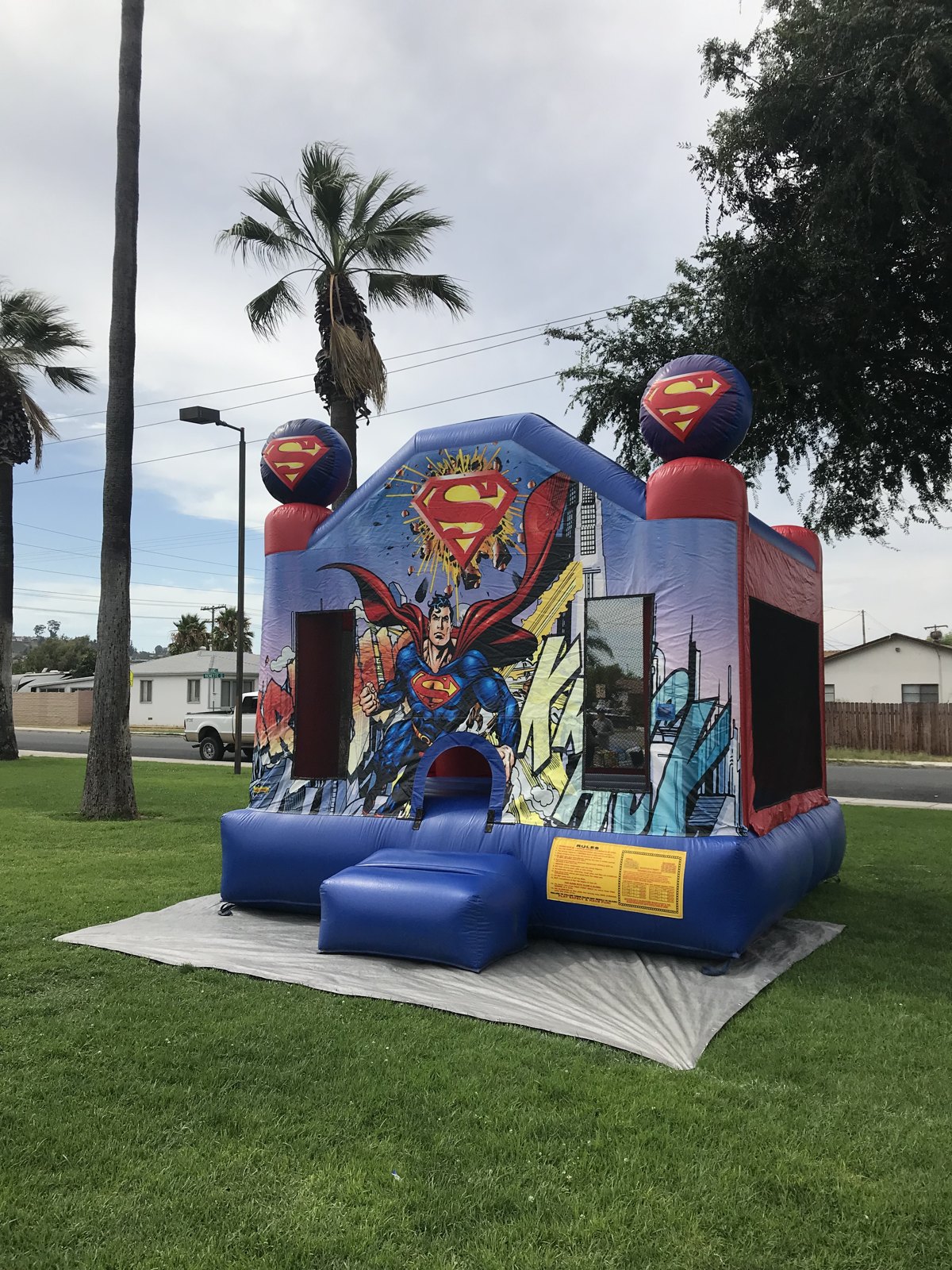 13x13 Superman bounce house rental in San Diego — angled front view on a protective tarp showing the entry ramp and rules panel