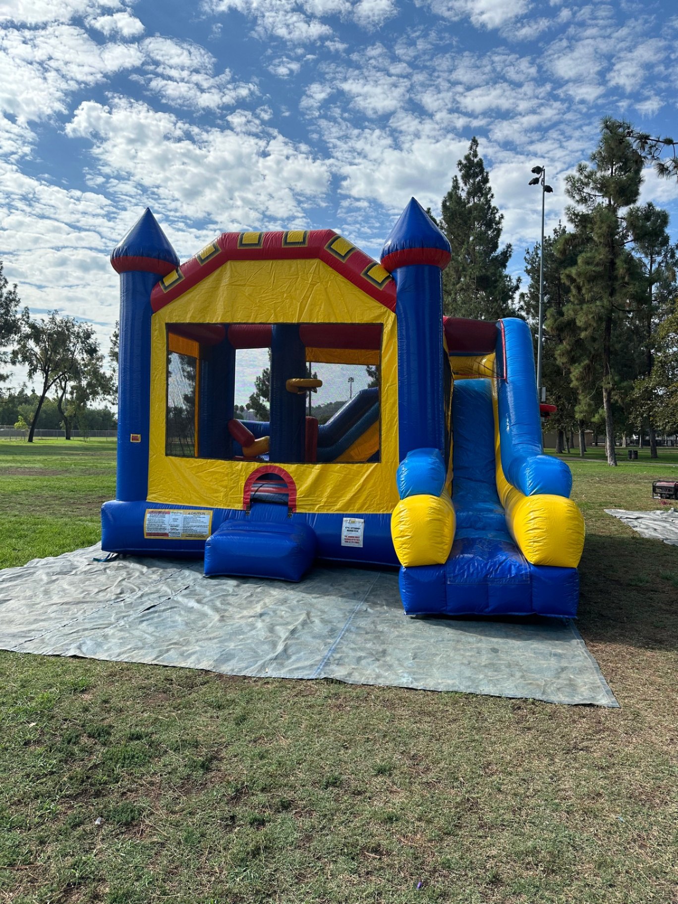 6n1 blue castle combo bounce house rental in San Diego, front view of the blue, yellow and red castle with side slide set up on a tarp at a park