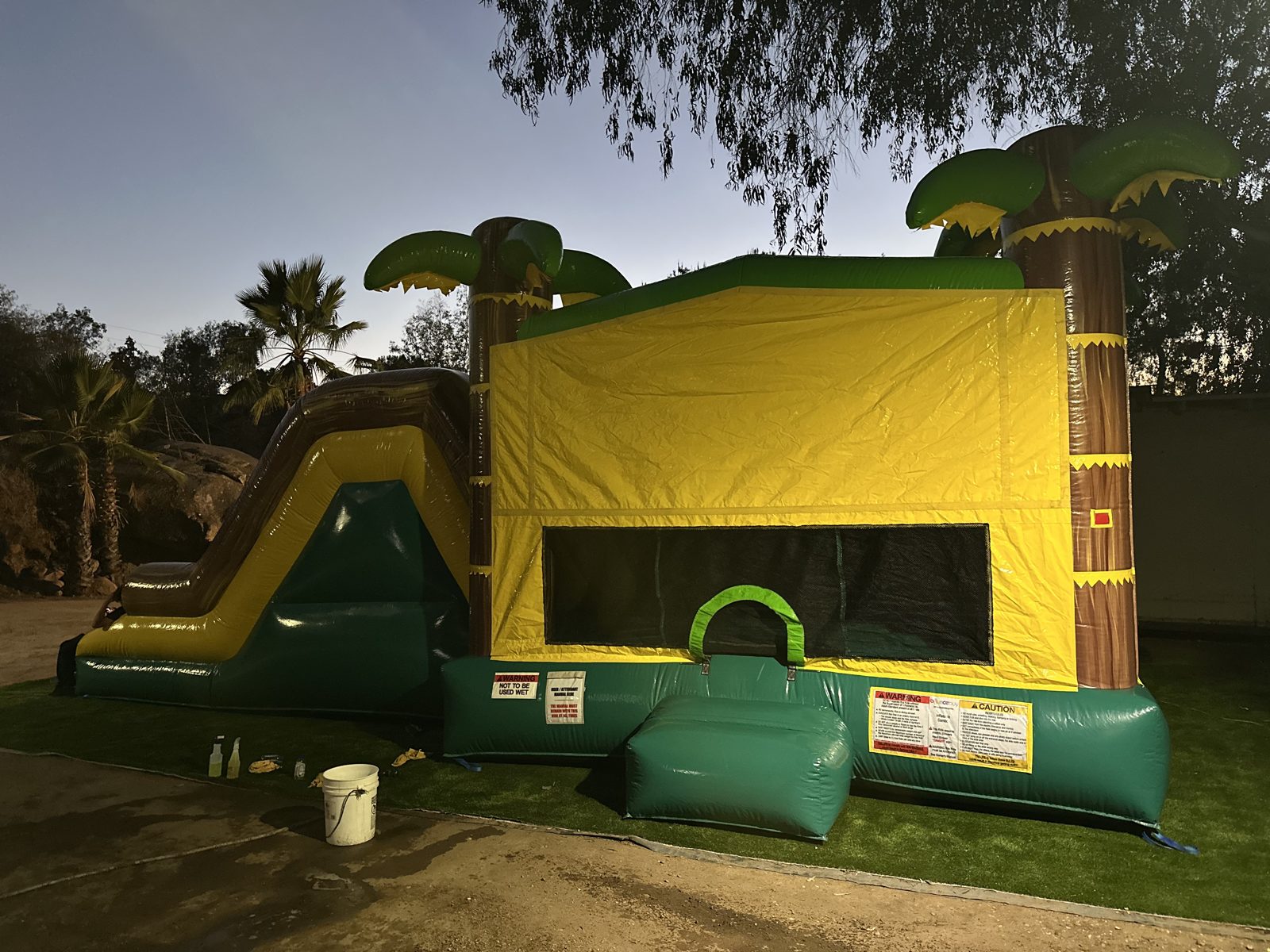 4n1 tropical oasis dual slide combo bounce house rental in San Diego, evening setup on turf with the entry and palm tree towers